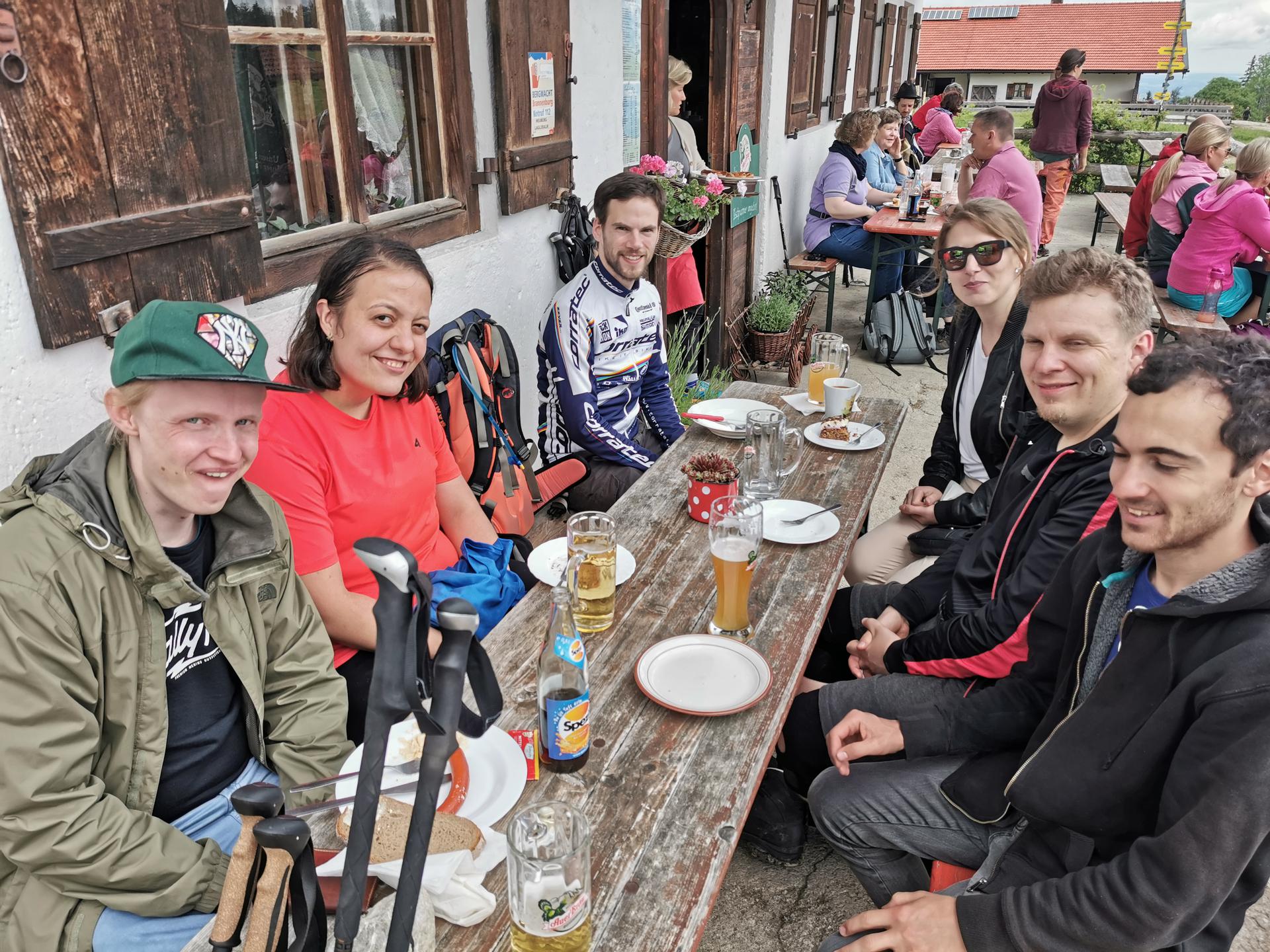 6 Personen aus der Gruppe sitzen an einer Biergarnitur vor der Lagler Hütte. Auf dem Tisch sind Teller mit Kuchen oder Brotzeit (teilweise bereits leer) und Bier, Limo und Heißgetränke zu sehen. Die Hütte ist gut besucht.