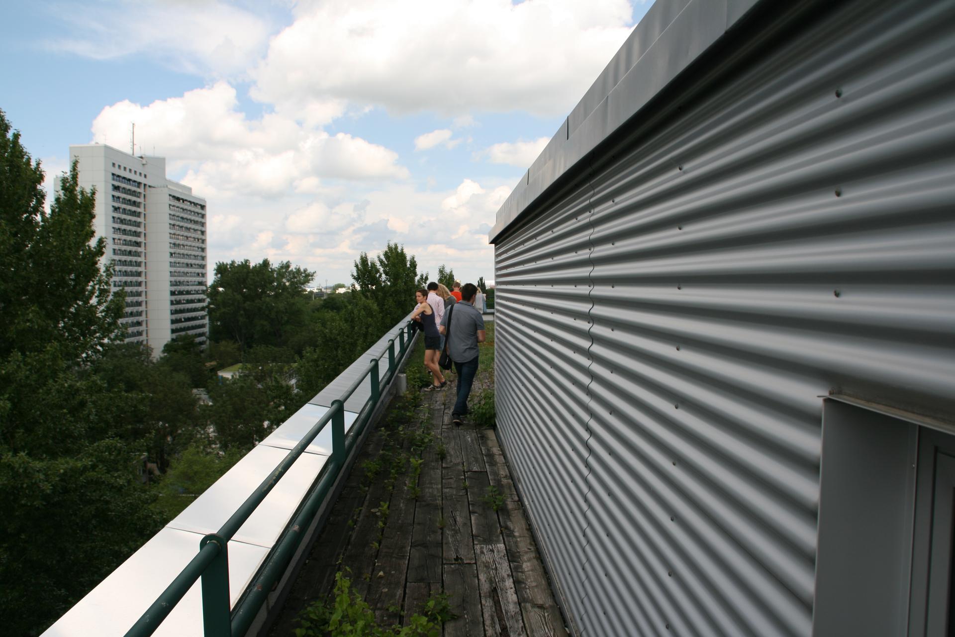 Ein etwa 1m breiter Weg aus Holzplanken führt am Wellblech verkleideten Aufbau auf die andere Seite des MKH-Daches. Im Hintergrund das HSH. Etwa 5 Personen.